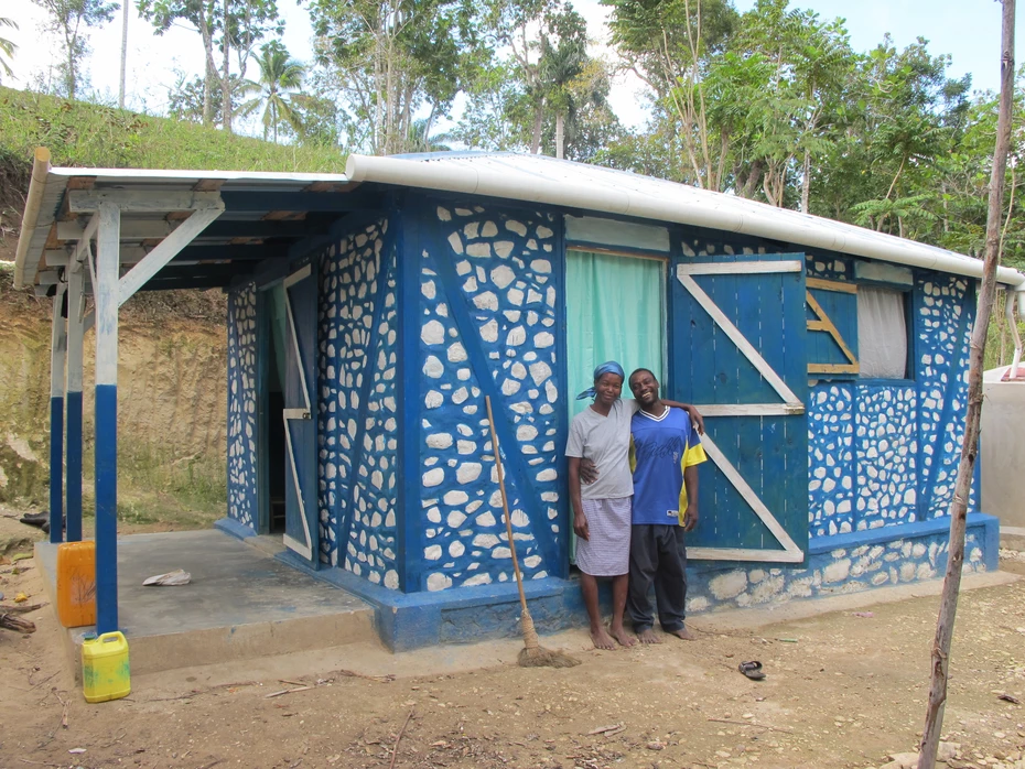 Famiglia felice davanti alla nuova casa Due persone sorridenti davanti a una piccola casa bianca e blu. La solida casa, costruita con materiali locali, offre protezione e sicurezza.