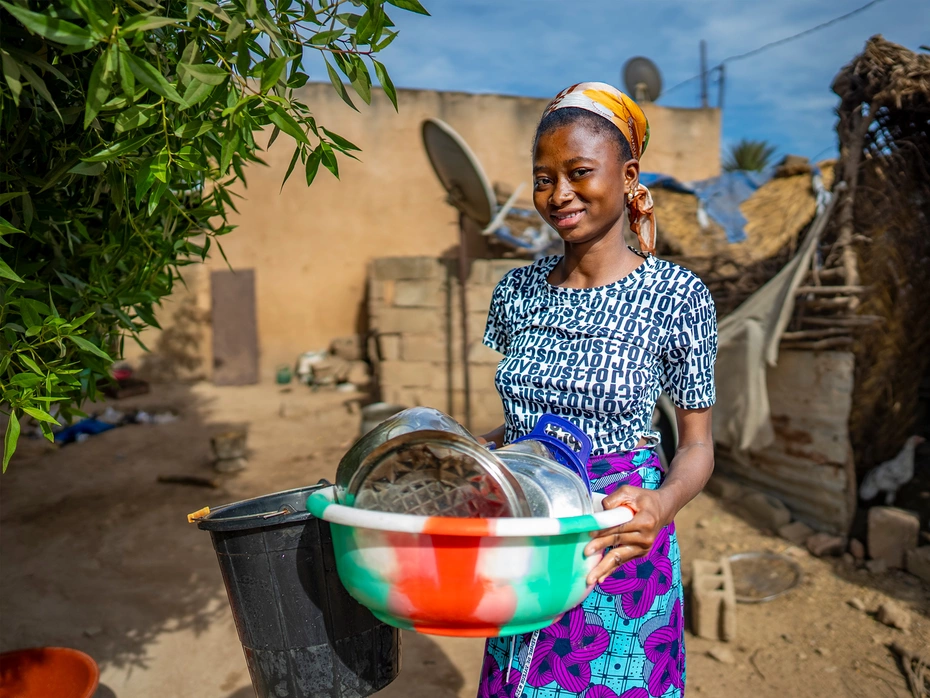 Una donna sorride con una grande cesta piena di stoviglie davanti a una capanna.