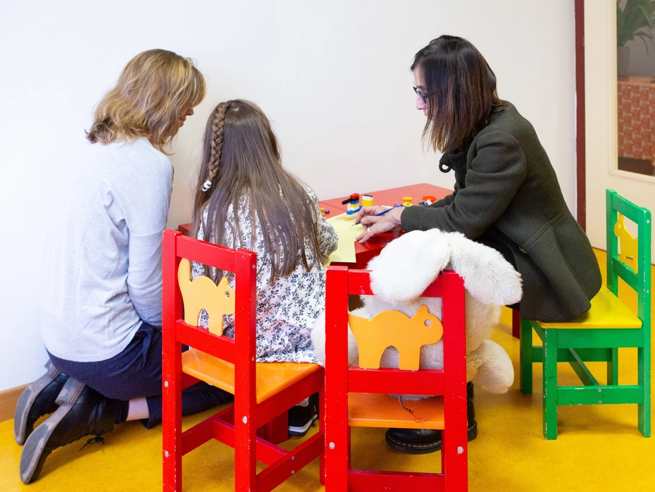 Una bambina e due donne adulte sedute a un tavolino, viste da dietro.