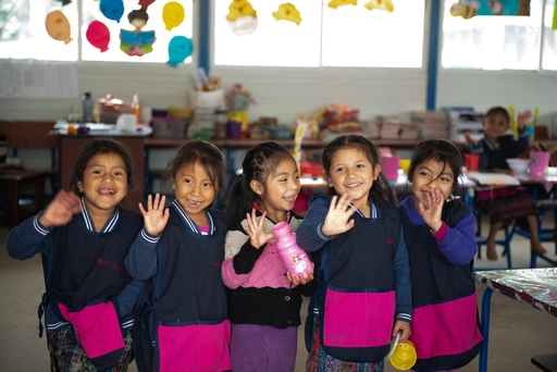 Cinque bambine vestite di blu e rosa sorridono felicemente e salutano la fotocamera nella loro classe.